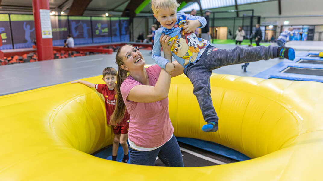 Eine Frau hebt einen lächelnden Jungen über einen gelben aufblasbaren Ring im JUMP DOME Klagenfurt, einem Indoor-Spielzentrum, während ein anderes Kind in der Nähe steht.