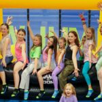 Eine Gruppe von Kindern in bunten Kleidern sitzt auf dem Rand einer Hüpfburg im JUMP DOME Klagenfurt, einem Indoor-Trampolinpark, wobei ein Kind auf der Matte davor sitzt.