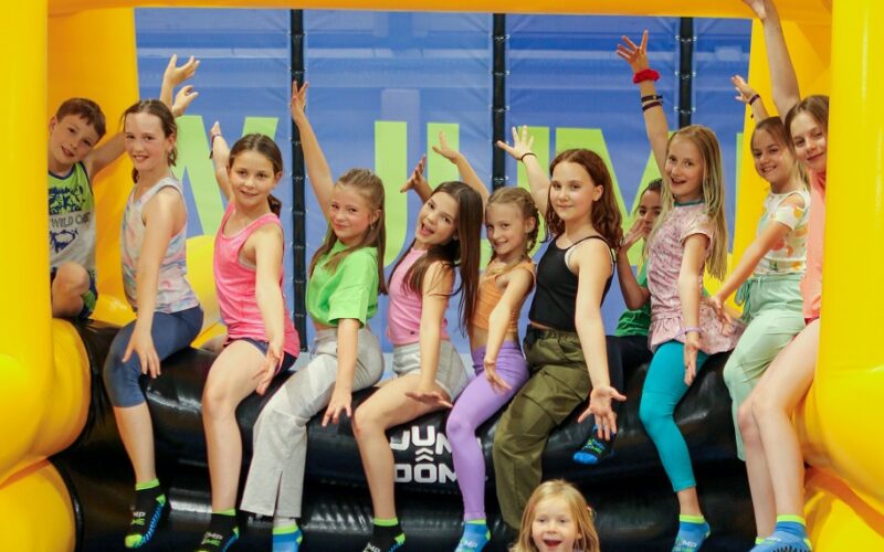 Eine Gruppe von Kindern in bunten Kleidern sitzt auf dem Rand einer Hüpfburg im JUMP DOME Klagenfurt, einem Indoor-Trampolinpark, wobei ein Kind auf der Matte davor sitzt.