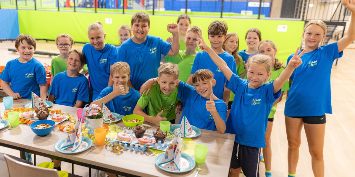 Eine Gruppe von Kindern in passenden blauen und grünen Hemden versammelt sich um einen Tisch mit Party-Essen und Dekoration, lächelt und posiert für ein Foto im JUMP DOME Klagenfurt, einem Indoor-Freizeitzentrum.