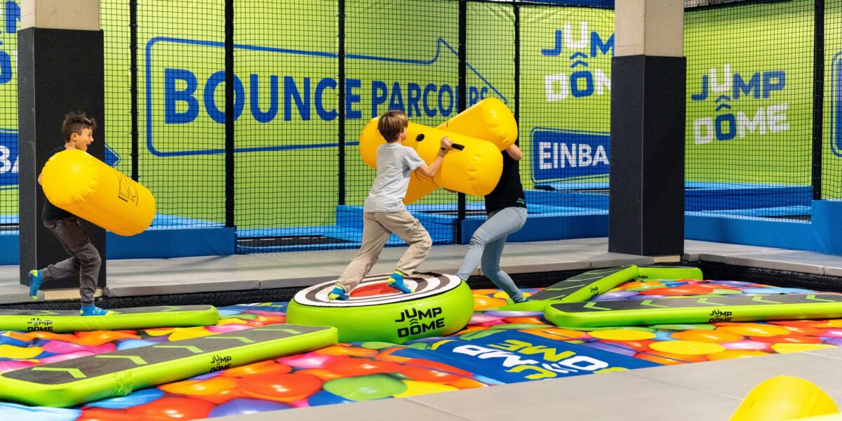 Drei Kinder spielen mit großen, gelben, gepolsterten Stöcken auf einer aufblasbaren Plattform in einem Indoor-Trampolin- und Aktivitätspark mit der Beschilderung „Bounce“ und „Jump Dome“.