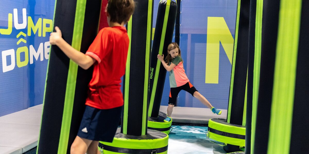 Zwei Kinder überwinden hängende Hindernisse in einem Indoor-Spielbereich mit gepolsterten Plattformen und einer blauen Wand mit der Aufschrift „JUMP DOME“.
