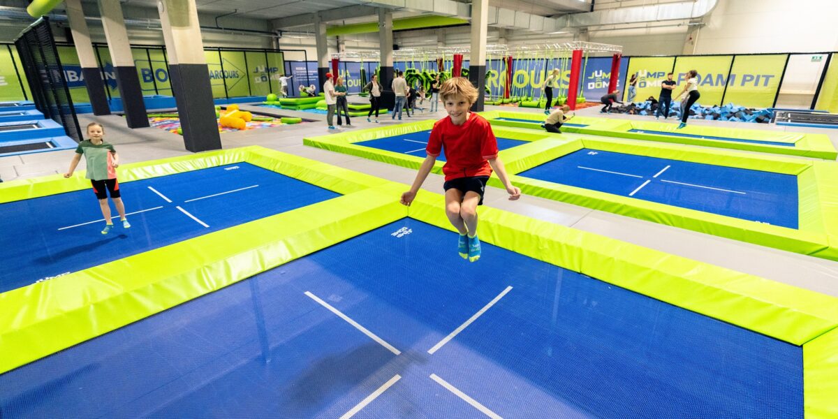 Kinder springen auf blauen und grünen Trampolinen in einem Indoor-Trampolinpark, im Hintergrund sind andere Personen und Geräte zu sehen.