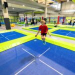 Kinder springen auf blauen und grünen Trampolinen in einem Indoor-Trampolinpark, im Hintergrund sind andere Personen und Geräte zu sehen.