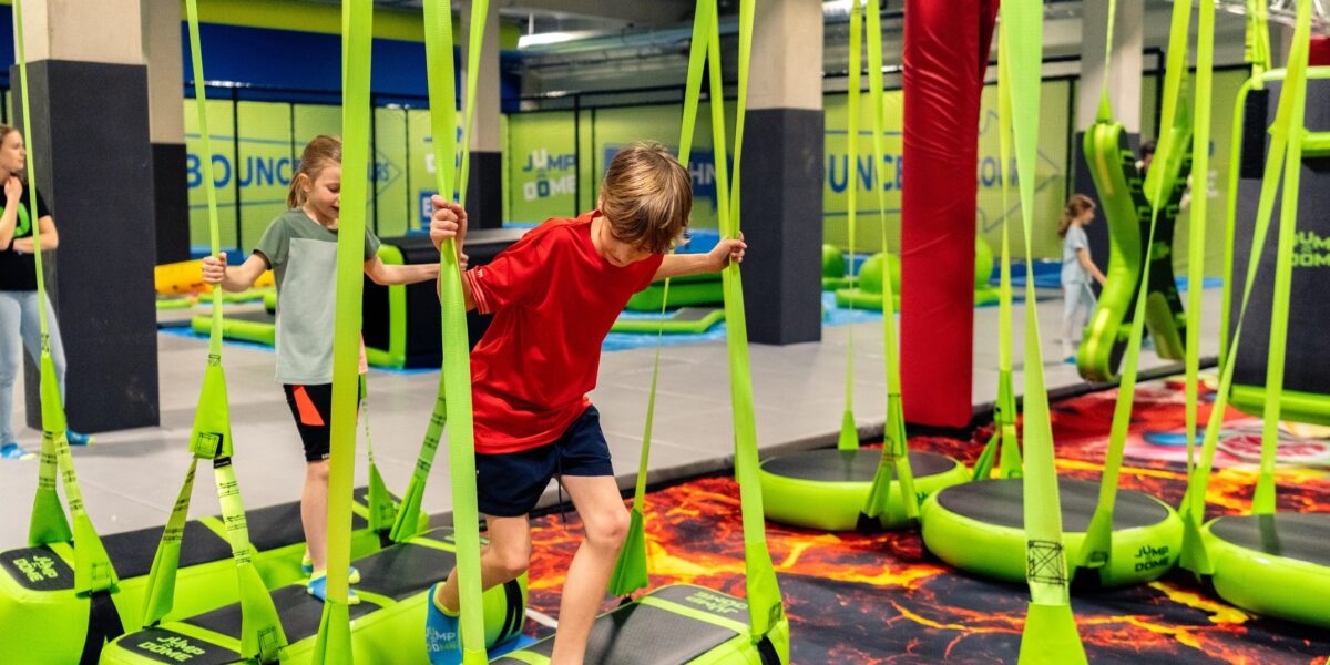 Kinder balancieren auf hängenden grünen Plattformen mit Gurten in einem Indoor-Trampolinpark, umgeben von weichen Polstern und Hindernisparcours-Elementen.