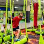 Kinder balancieren auf hängenden grünen Plattformen mit Gurten in einem Indoor-Trampolinpark, umgeben von weichen Polstern und Hindernisparcours-Elementen.