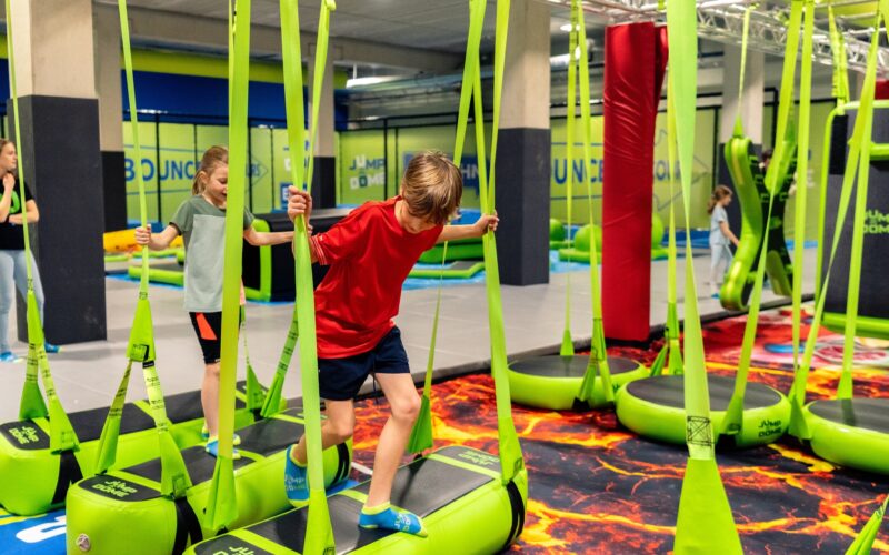 Kinder balancieren auf hängenden grünen Plattformen mit Gurten in einem Indoor-Trampolinpark, umgeben von weichen Polstern und Hindernisparcours-Elementen.