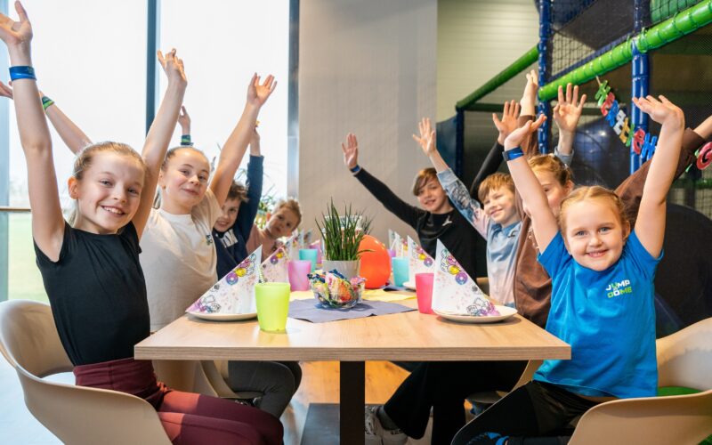 Eine Gruppe Kinder sitzt mit Partyhüten und -bechern um einen dekorierten Tisch, alle lächeln und heben feierlich die Arme.