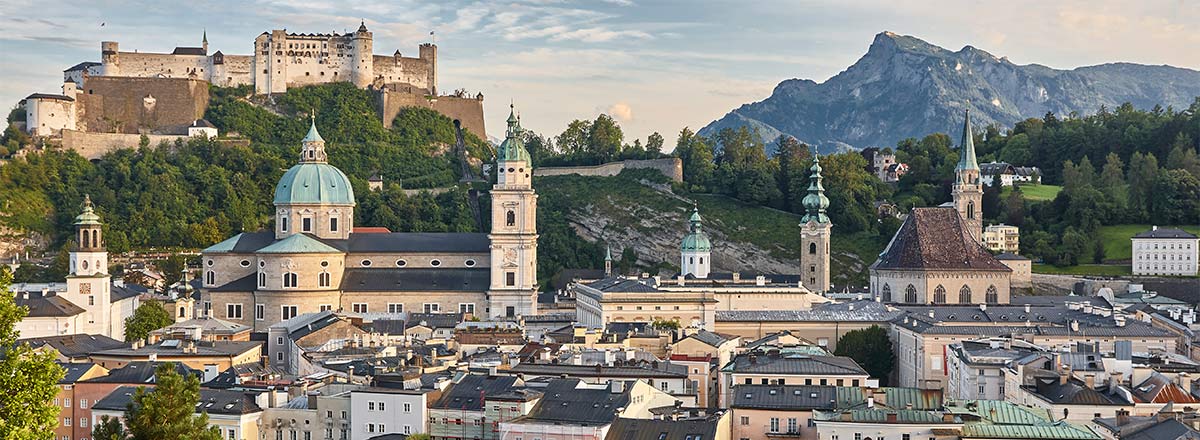Panoramablick auf Salzburg, Österreich, mit der Festung Hohensalzburg auf einem Hügel, historischen Gebäuden, Kirchenkuppeln und Bergen im Hintergrund.