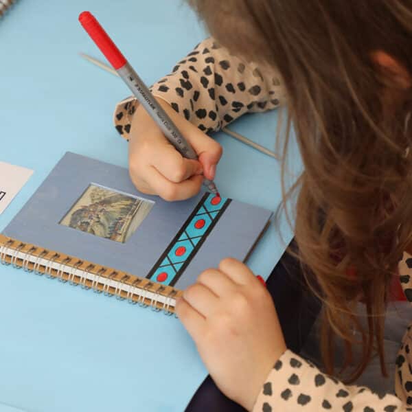 Ein Kind zeichnet mit einem roten Stift geometrische Muster in ein Notizbuch und verwendet dabei ein gedrucktes Design als Vorlage. Farbige Marker und ein weiteres Notizbuch liegen in der Nähe auf dem Tisch.