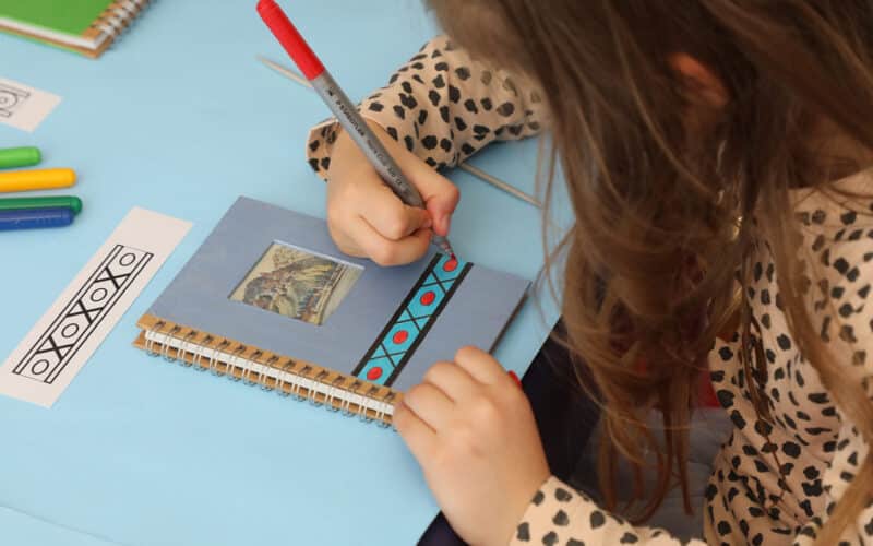 Ein Kind zeichnet mit einem roten Stift geometrische Muster in ein Notizbuch und verwendet dabei ein gedrucktes Design als Vorlage. Farbige Marker und ein weiteres Notizbuch liegen in der Nähe auf dem Tisch.