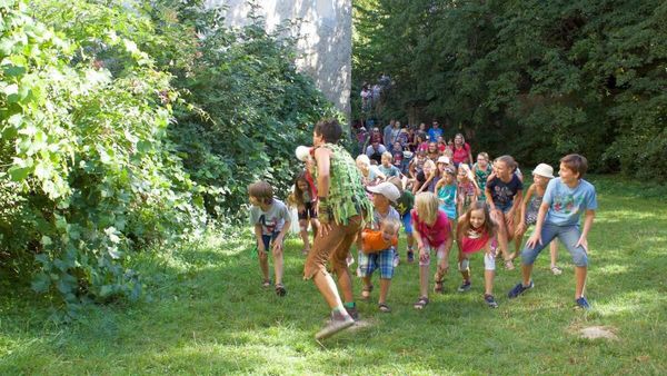 Eine Gruppe Kinder folgt einem Erwachsenen in einem grünen Kostüm durch eine Rasenfläche mit Bäumen und einem Gebäude im Hintergrund.