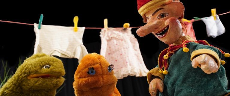 Vor einem dunklen Hintergrund stehen drei bunte Puppen vor einer Wäscheleine, an der Kinderkleidung hängt.