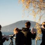 Vier Personen stehen mit Ferngläsern, Notizbüchern und einem Teleskop in der Nähe eines Seeufers und beobachten die Umgebung unter einem Baum mit Herbstlaub.