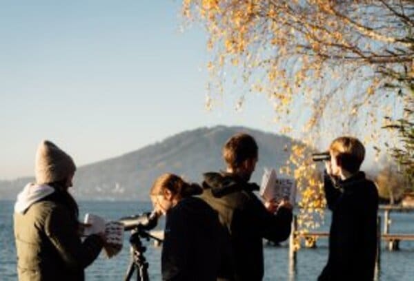 Vier Personen stehen mit Ferngläsern, Notizbüchern und einem Teleskop in der Nähe eines Seeufers und beobachten die Umgebung unter einem Baum mit Herbstlaub.