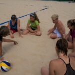 Eine Gruppe junger Leute sitzt im Kreis auf einem Beachvolleyballfeld und hört einem Trainer zu, der mit dem Finger in den Sand zeichnet. Mehrere Volleybälle liegen auf dem Boden.