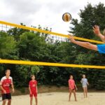Fünf Kinder spielen Volleyball auf einem Sandplatz im Freien. Ein Spieler in einem orangefarbenen Trikot springt, um den Ball über das Netz zu schlagen. Im Hintergrund sind Bäume und ein bewölkter Himmel zu sehen.