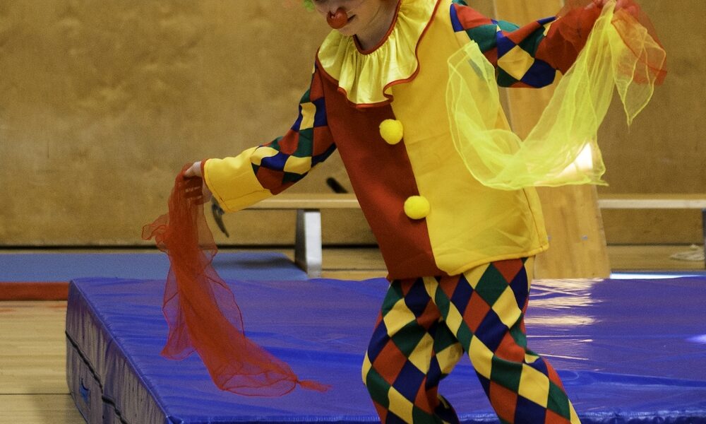 Eine als Clown verkleidete Person balanciert auf einem rollenden Zylinder und hält dabei bunte Tücher in einer Turnhalle.