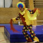 Eine als Clown verkleidete Person balanciert auf einem rollenden Zylinder und hält dabei bunte Tücher in einer Turnhalle.