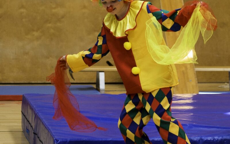 Eine als Clown verkleidete Person balanciert auf einem rollenden Zylinder und hält dabei bunte Tücher in einer Turnhalle.