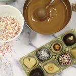 Ein Tablett mit verschiedenen Pralinen, eine Schüssel geschmolzene Schokolade mit einem Löffel, eine Tasse mit bunten Streuseln und Schokoladenraspeln auf einer Marmoroberfläche.