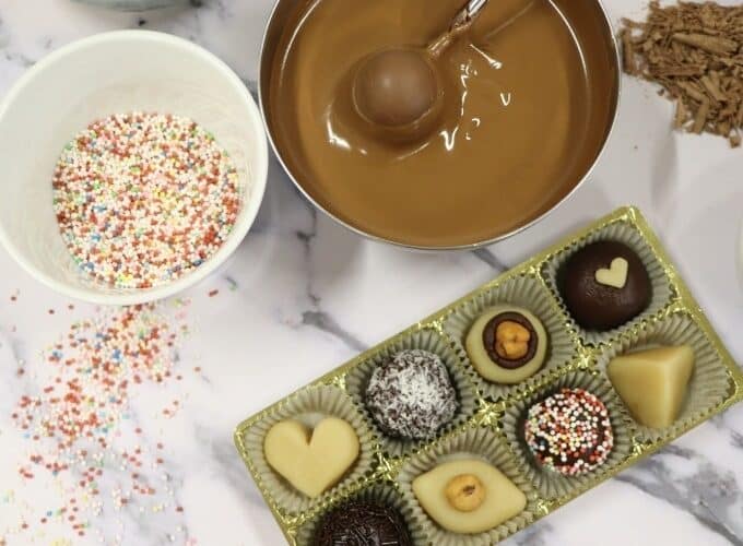 Ein Tablett mit verschiedenen Pralinen, eine Schüssel geschmolzene Schokolade mit einem Löffel, eine Tasse mit bunten Streuseln und Schokoladenraspeln auf einer Marmoroberfläche.