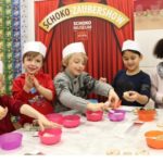 Fünf Kinder mit Papierhüten stellen an einem Tisch im Schoko-Museum Schokoladenleckereien her. Im Hintergrund stehen Schüsseln mit Zutaten und eine Schautafel mit der Aufschrift „Schoko-Zaubershow“.