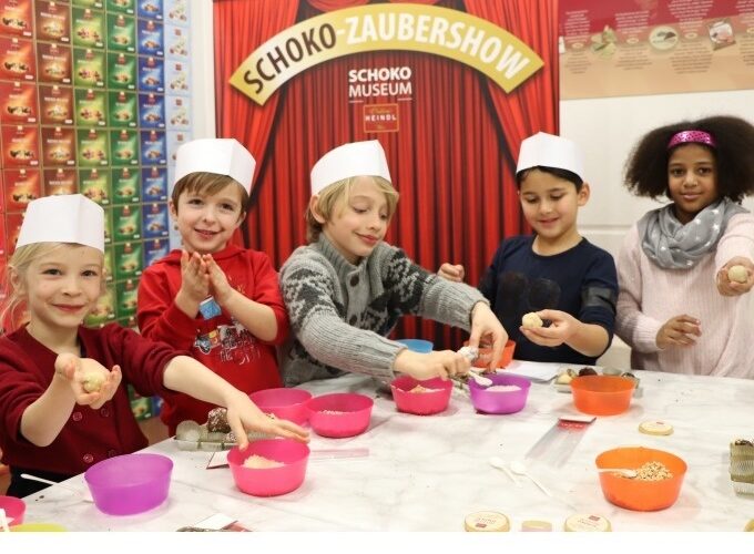 Fünf Kinder mit Papierhüten stellen an einem Tisch im Schoko-Museum Schokoladenleckereien her. Im Hintergrund stehen Schüsseln mit Zutaten und eine Schautafel mit der Aufschrift „Schoko-Zaubershow“.