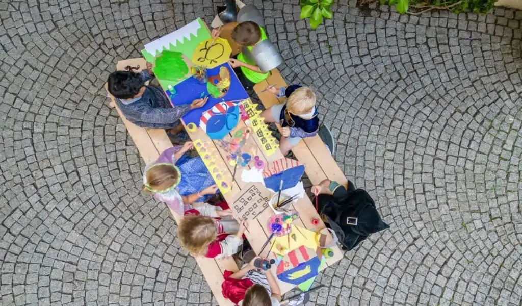 Eine Gruppe von Kindern sitzt um einen Holztisch im Freien und malt und zeichnet auf buntem Papier, das von oben auf einem Kopfsteinpflaster liegt.
