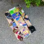 Eine Gruppe von Kindern sitzt um einen Holztisch im Freien und malt und zeichnet auf buntem Papier, das von oben auf einem Kopfsteinpflaster liegt.
