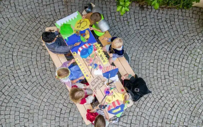 Eine Gruppe von Kindern sitzt um einen Holztisch im Freien und malt und zeichnet auf buntem Papier, das von oben auf einem Kopfsteinpflaster liegt.