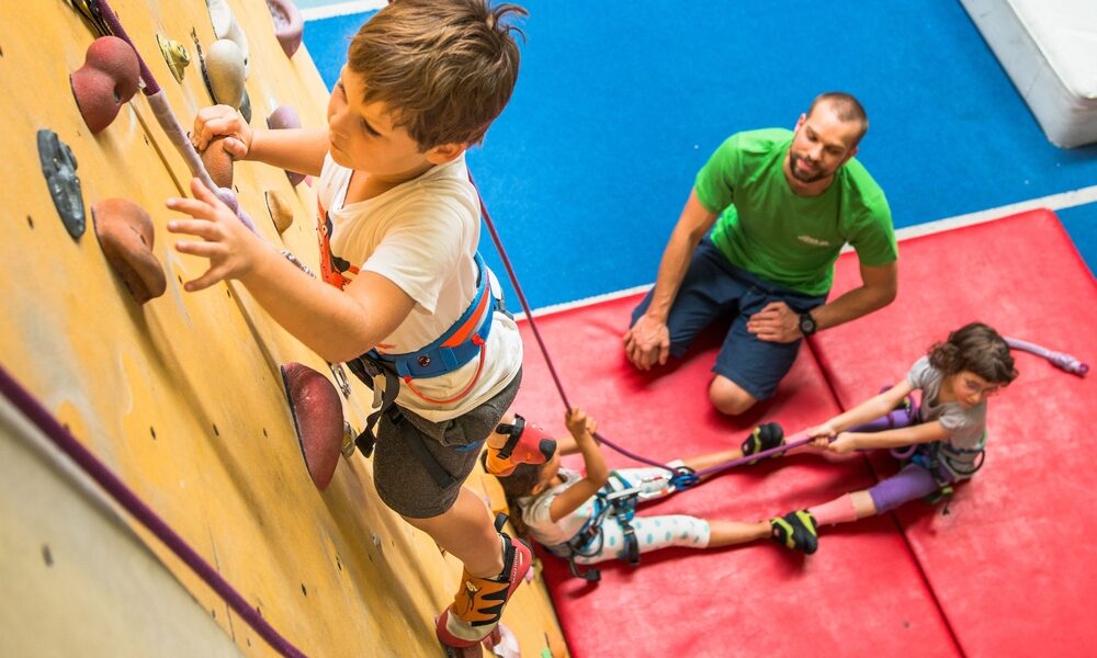 Ein kleiner Junge klettert an einer Indoor-Felswand, während zwei Kinder sein Sicherungsseil halten und ein Erwachsener vom Boden aus auf einer blau-roten Matte die Aufsicht führt.