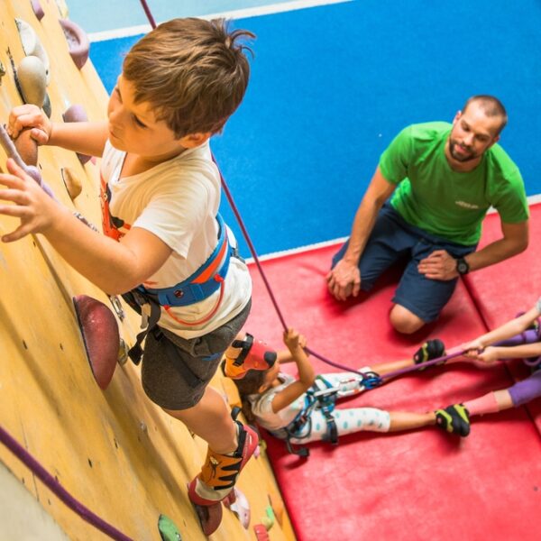 Ein kleiner Junge klettert an einer Indoor-Felswand, während zwei Kinder sein Sicherungsseil halten und ein Erwachsener vom Boden aus auf einer blau-roten Matte die Aufsicht führt.