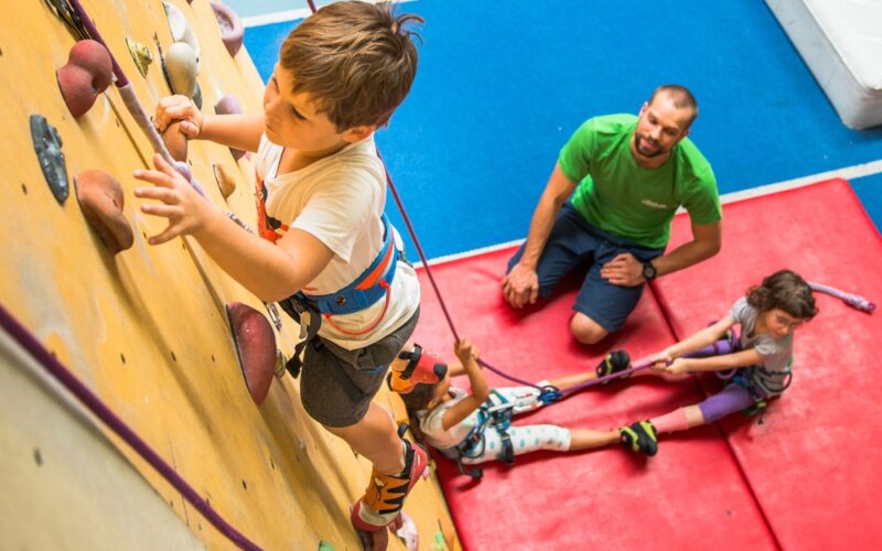 Ein kleiner Junge klettert an einer Indoor-Felswand, während zwei Kinder sein Sicherungsseil halten und ein Erwachsener vom Boden aus auf einer blau-roten Matte die Aufsicht führt.
