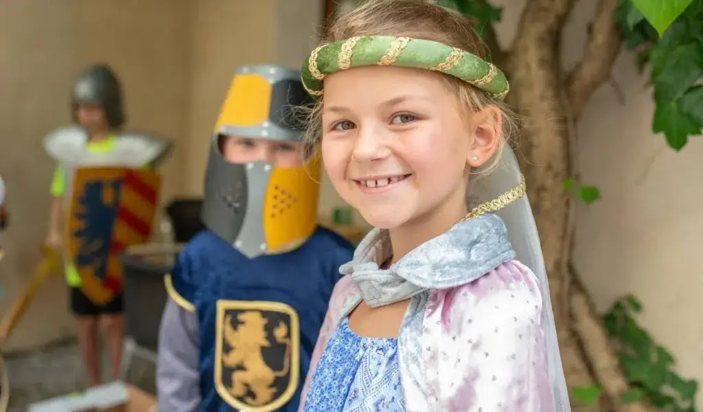 Kinder in mittelalterlichen Kostümen, mit einem lächelnden Mädchen im Vordergrund und einem Jungen in einem Ritterkostüm hinter ihr.