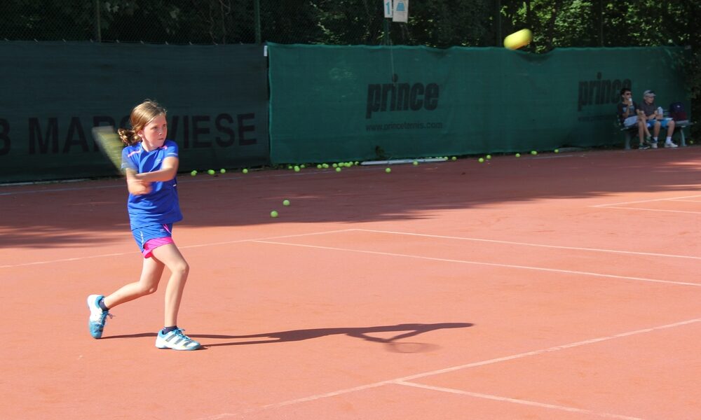 Ein junges Mädchen in blauer Sportkleidung schwingt einen Tennisschläger auf einem Sandplatz im Freien, während im Hintergrund mehrere Tennisbälle verstreut sind.