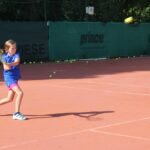Ein junges Mädchen in blauer Sportkleidung schwingt einen Tennisschläger auf einem Sandplatz im Freien, während im Hintergrund mehrere Tennisbälle verstreut sind.