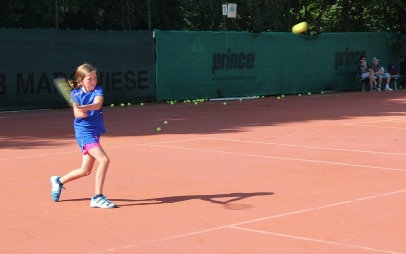 Ein junges Mädchen in blauer Sportkleidung schwingt einen Tennisschläger auf einem Sandplatz im Freien, während im Hintergrund mehrere Tennisbälle verstreut sind.