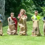 Sechs Kinder stehen mit Sackleinen im Gras und scheinen bereit zu sein, ein Sackhüpfen zu veranstalten, während im Hintergrund Bäume und eine Steinmauer zu sehen sind.