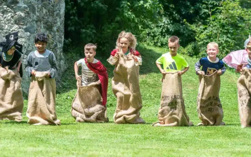Sechs Kinder stehen mit Sackleinen im Gras und scheinen bereit zu sein, ein Sackhüpfen zu veranstalten, während im Hintergrund Bäume und eine Steinmauer zu sehen sind.