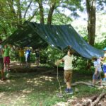 Eine Gruppe von Kindern und ein Erwachsener bauen an einem sonnigen Tag in einem Waldgebiet eine große grüne Plane als Unterschlupf auf.