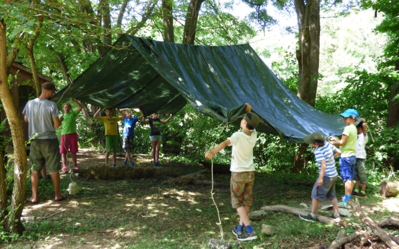 Eine Gruppe von Kindern und ein Erwachsener bauen an einem sonnigen Tag in einem Waldgebiet eine große grüne Plane als Unterschlupf auf.