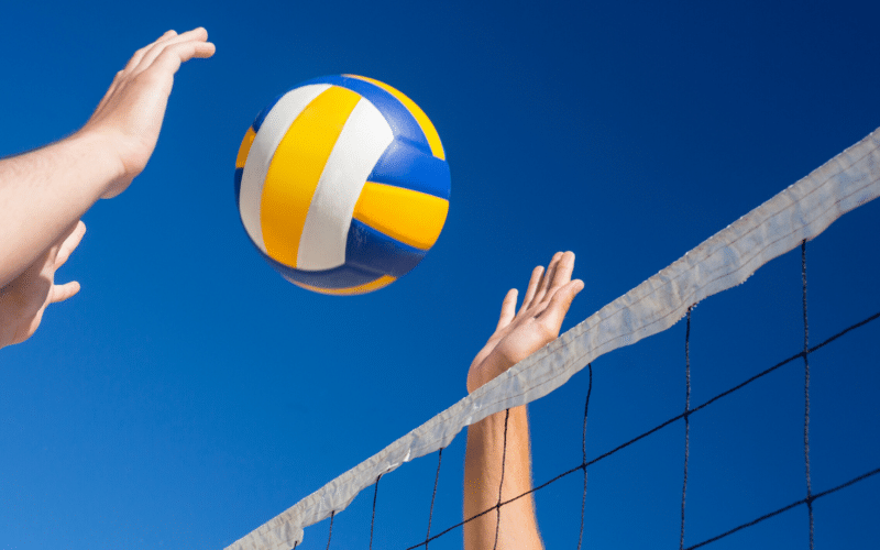 Ein Volleyball schwebt in der Luft über dem Netz, während zwei Spieler vor einem klaren blauen Himmel danach greifen.