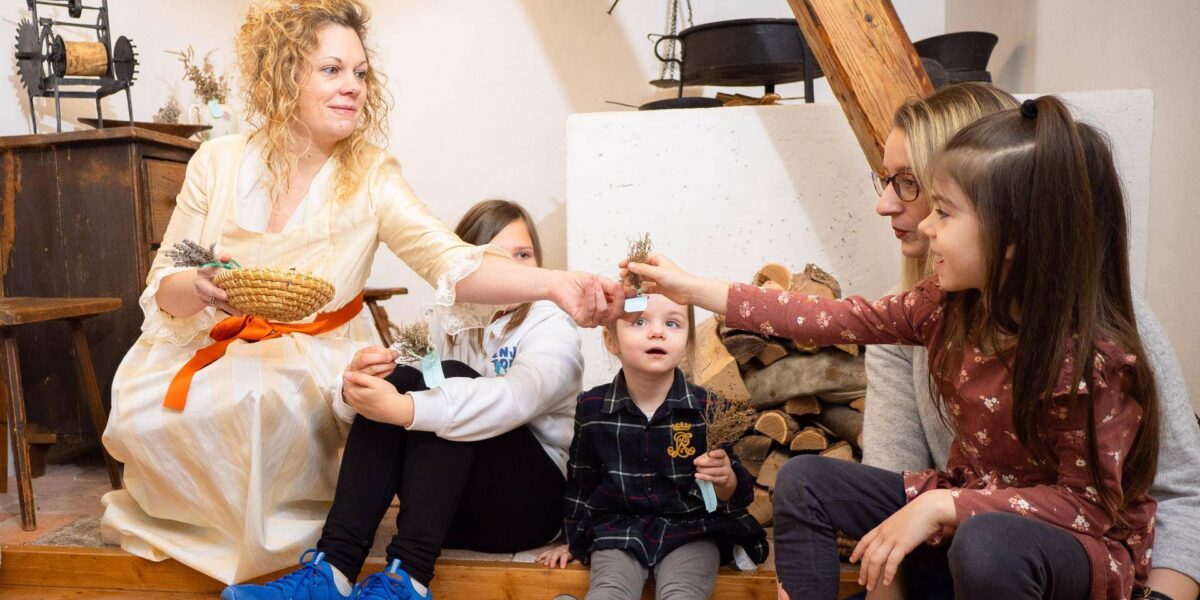 Eine Frau in einem weißen Kleid überreicht einem Mädchen, das mit anderen Kindern und einem Erwachsenen in einem rustikalen Raum auf dem Boden sitzt, einen kleinen Gegenstand.