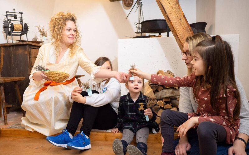 Eine Frau in einem weißen Kleid überreicht einem Mädchen, das mit anderen Kindern und einem Erwachsenen in einem rustikalen Raum auf dem Boden sitzt, einen kleinen Gegenstand.