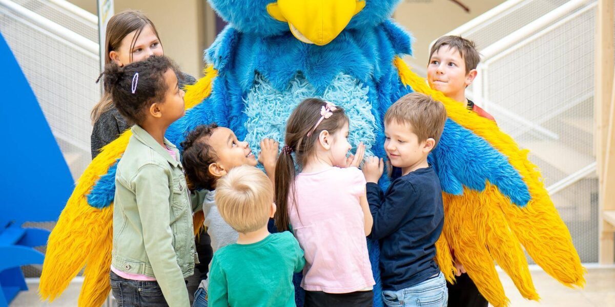 In einem Gebäude steht eine Gruppe Kinder herum und umarmt eine Person in einem großen, bunten blau-gelben Vogelkostüm.