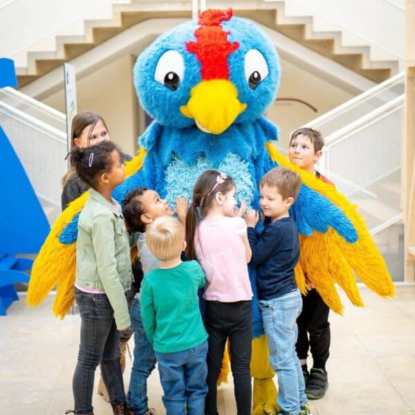 In einem Gebäude steht eine Gruppe Kinder herum und umarmt eine Person in einem großen, bunten blau-gelben Vogelkostüm.