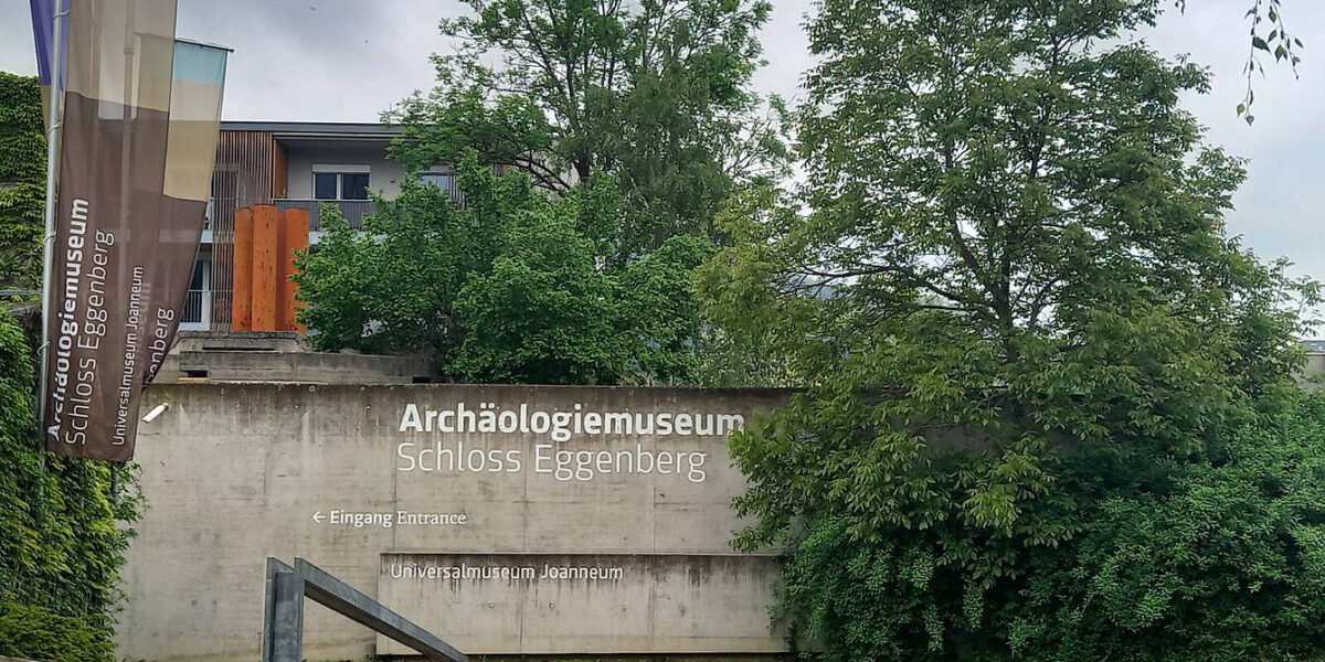 Betonwand mit Beschilderung „Archäologiemuseum Schloss Eggenberg“, Eingangsschild, Bannern, Bäumen und im Hintergrund ist ein Teil eines modernen Gebäudes zu sehen.