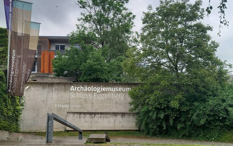 Betonwand mit Beschilderung „Archäologiemuseum Schloss Eggenberg“, Eingangsschild, Bannern, Bäumen und im Hintergrund ist ein Teil eines modernen Gebäudes zu sehen.