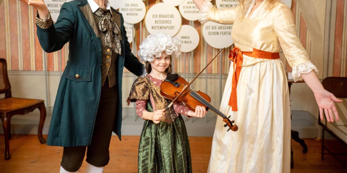 Drei Personen in historischen Kostümen, ein Kind mit einer Geige, posieren und lächeln drinnen vor einer Wand mit deutschen Sprechblasen.